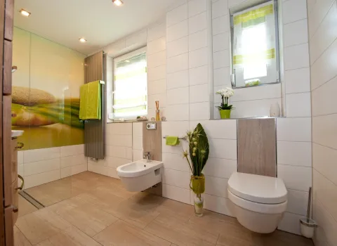 Helles, modernes Badezimmer mit beiger Bodenfliese, wandhängendem WC und Bidet, dekorativen Pflanzen und grünen Akzenten. Im Hintergrund eine Dusche mit Glaswand und Naturmotiv.